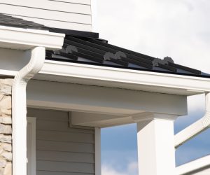 Close-up of a house roof with black metal panels, white gutters, stone siding, and white trim.