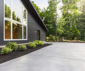 Modern dark grey house with large windows, concrete driveway, and landscaped shrubs, surrounded by trees.