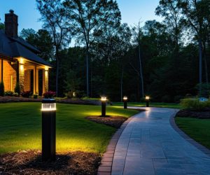 Light posts illuminated backyard garden during night hours. Modern backyard outdoor lighting systems. A curved walkway lined with illuminated bollard lights leads to a house at dusk, surrounded by trees and landscaped lawn.